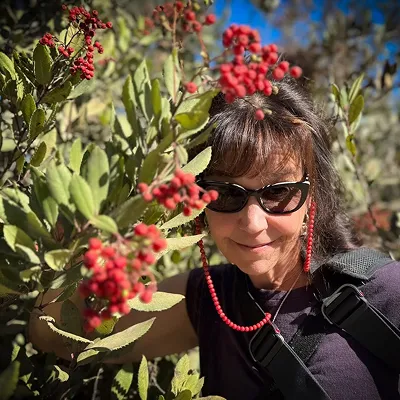 A woman wearing sunglasses and a dark shirt stands among green foliage and clusters of red berries, with sunlight casting shadows on her face. She is partially hidden by the branches.