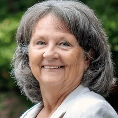 An older woman with gray hair and a light-colored blazer smiles outdoors in front of a blurred green background.