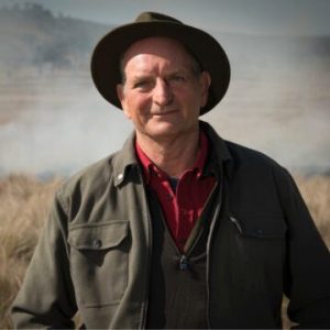 An older man wearing a wide-brimmed hat, red shirt, and dark jacket stands outdoors in a grassy field with a misty background and distant trees.