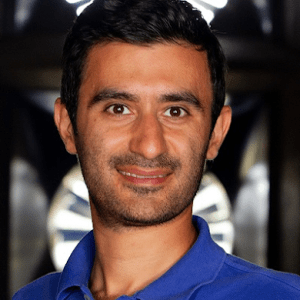 A man with short dark hair and a trimmed beard, wearing a blue polo shirt, stands in front of a blurred background with circular light patterns. He is looking at the camera and smiling slightly.