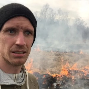 A man wearing a black beanie and brown hoodie looks concerned while standing in front of a field on fire, with flames and smoke rising in the background.