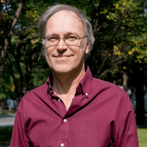 A middle-aged man with glasses and thinning hair, wearing a maroon button-up shirt, stands outdoors in a sunlit park with green trees in the background.