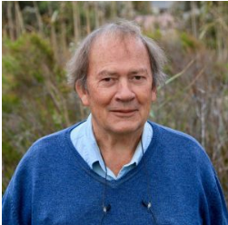 An older man with thinning gray hair, wearing a blue sweater over a light blue collared shirt, stands outdoors in front of tall grass and greenery, smiling gently at the camera.