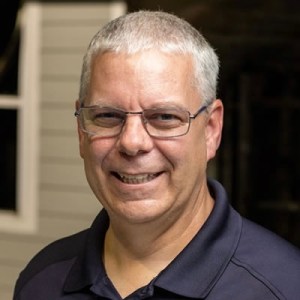 A middle-aged man with short gray hair and glasses smiles at the camera. He is wearing a navy blue polo shirt and standing indoors, with a window and siding visible in the background.