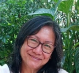 A woman with long dark hair and glasses smiles at the camera, with lush green plants in the background.