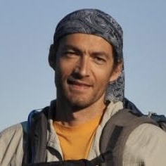 A man wearing a patterned bandana on his head, a light jacket, and a yellow shirt, standing outdoors with a backpack and smiling slightly against a clear blue sky.