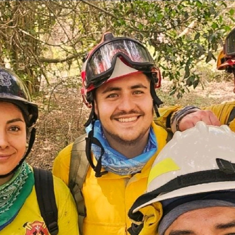 Three people wearing yellow firefighting uniforms, helmets, and green bandanas smile at the camera outdoors. Trees and greenery are visible in the background.