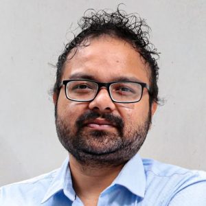 A man with dark curly hair, glasses, and a beard is wearing a light blue collared shirt and is standing against a plain, light-colored background.