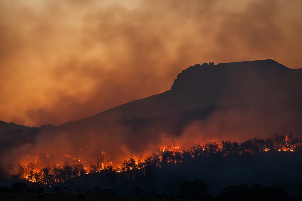 A wildfire burns through a forested hillside at dusk, sending thick smoke into the sky with an orange glow. Dark mountain silhouettes appear in the background, highlighting the intensity of the fire.