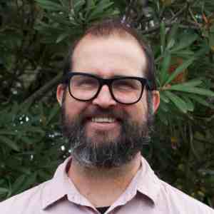 A bearded man with glasses and a light pink shirt smiles in front of leafy green foliage.