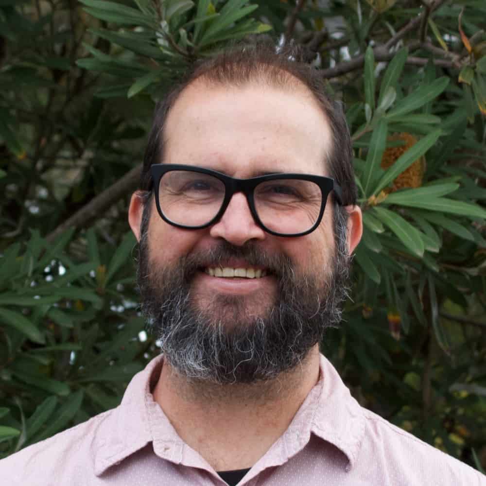 A bearded man with glasses and a light pink shirt smiles in front of leafy green foliage.