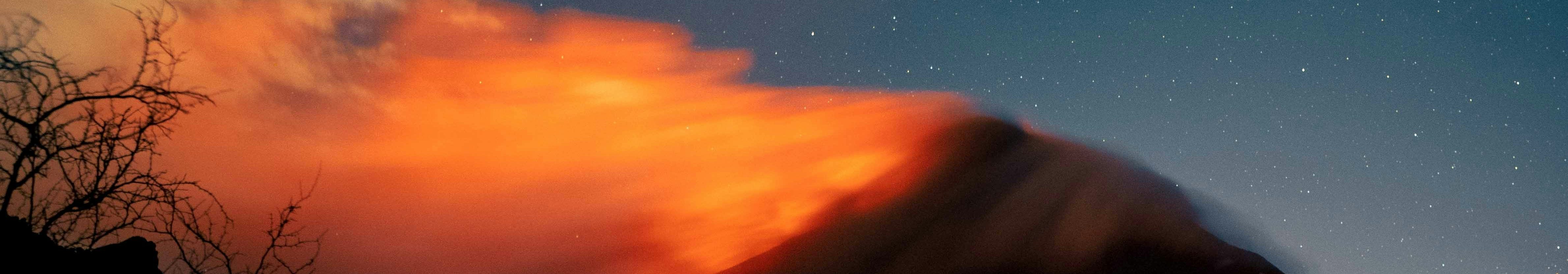 A wildfire burns at night, glowing orange and emitting a cloud of smoke, set against a starry sky. Silhouetted terrain and bare branches appear in the foreground.