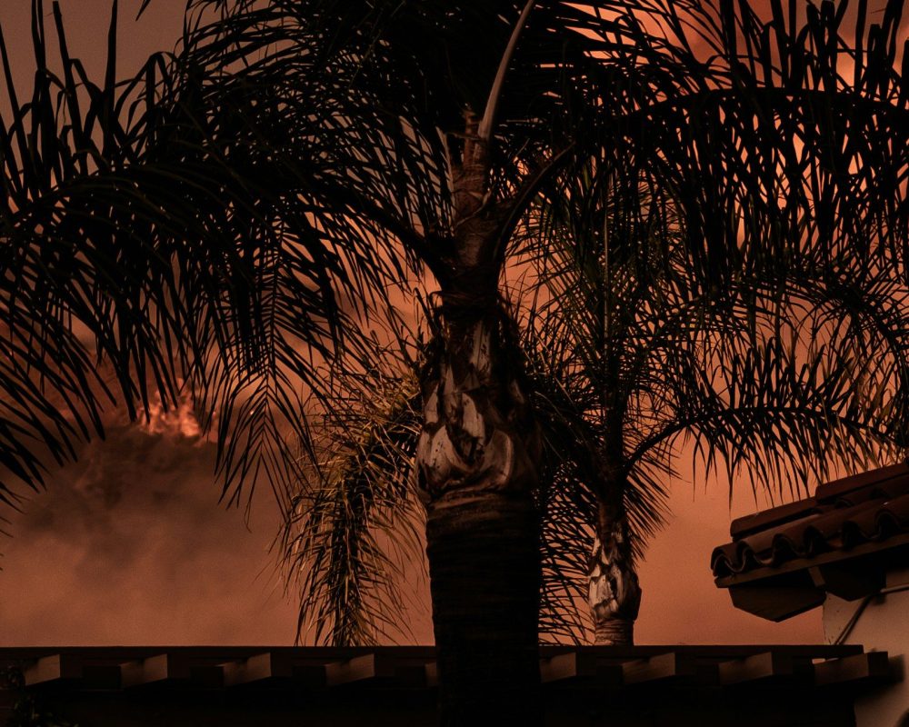 A large plume of orange smoke rises behind a house with a tiled roof and a palm tree, suggesting a wildfire burning near a mountain at dusk or night.