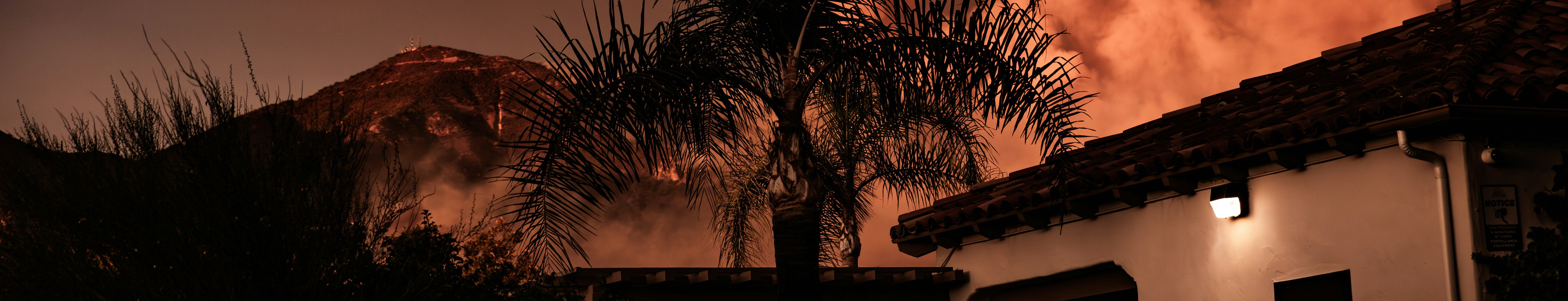A large plume of orange smoke rises behind a house with a tiled roof and a palm tree, suggesting a wildfire burning near a mountain at dusk or night.