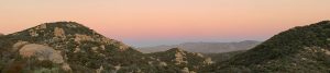 A serene landscape at sunset shows rolling green hills and rocky outcrops under a pastel sky with soft pink, orange, and blue hues. The foreground is filled with dense shrubs and vegetation.