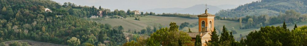 A church with a bell tower stands among dense green trees and rolling hills, with mountains in the background under a cloudy sky.