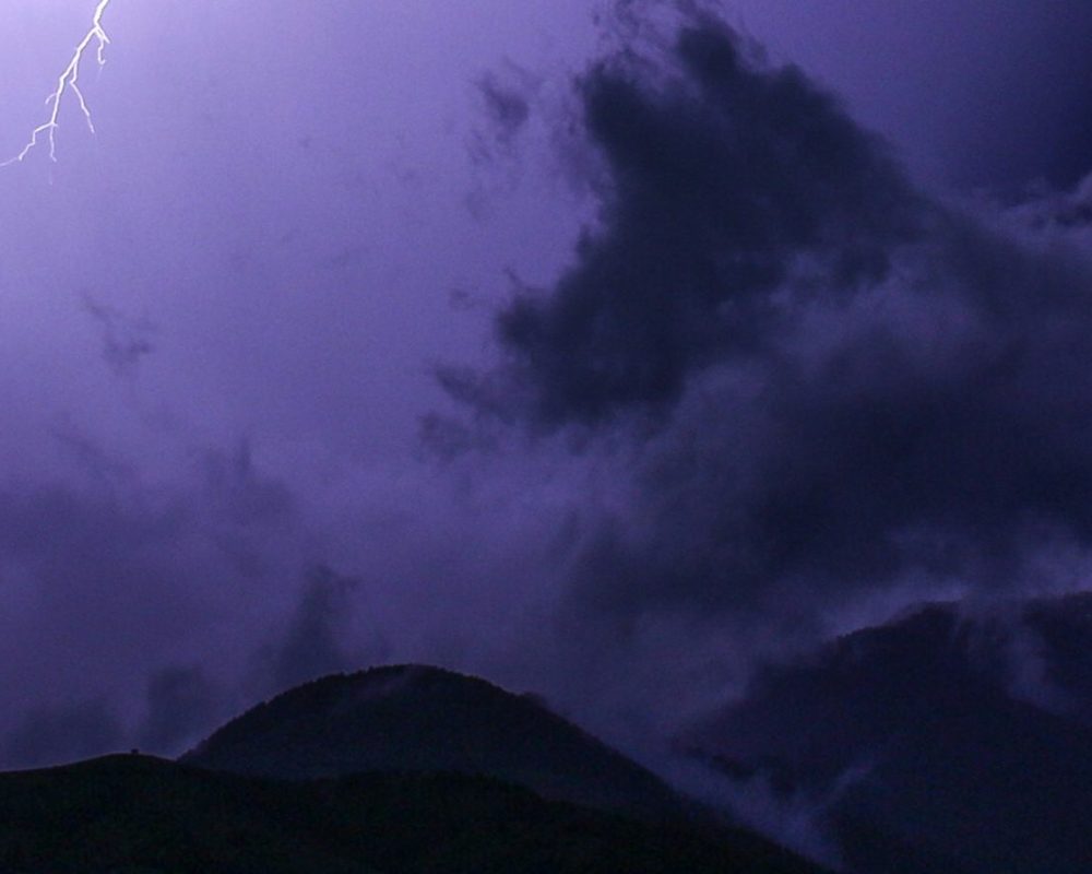 A jagged bolt of lightning illuminates dark clouds over silhouetted mountains at night, casting a purple-blue hue across the sky.