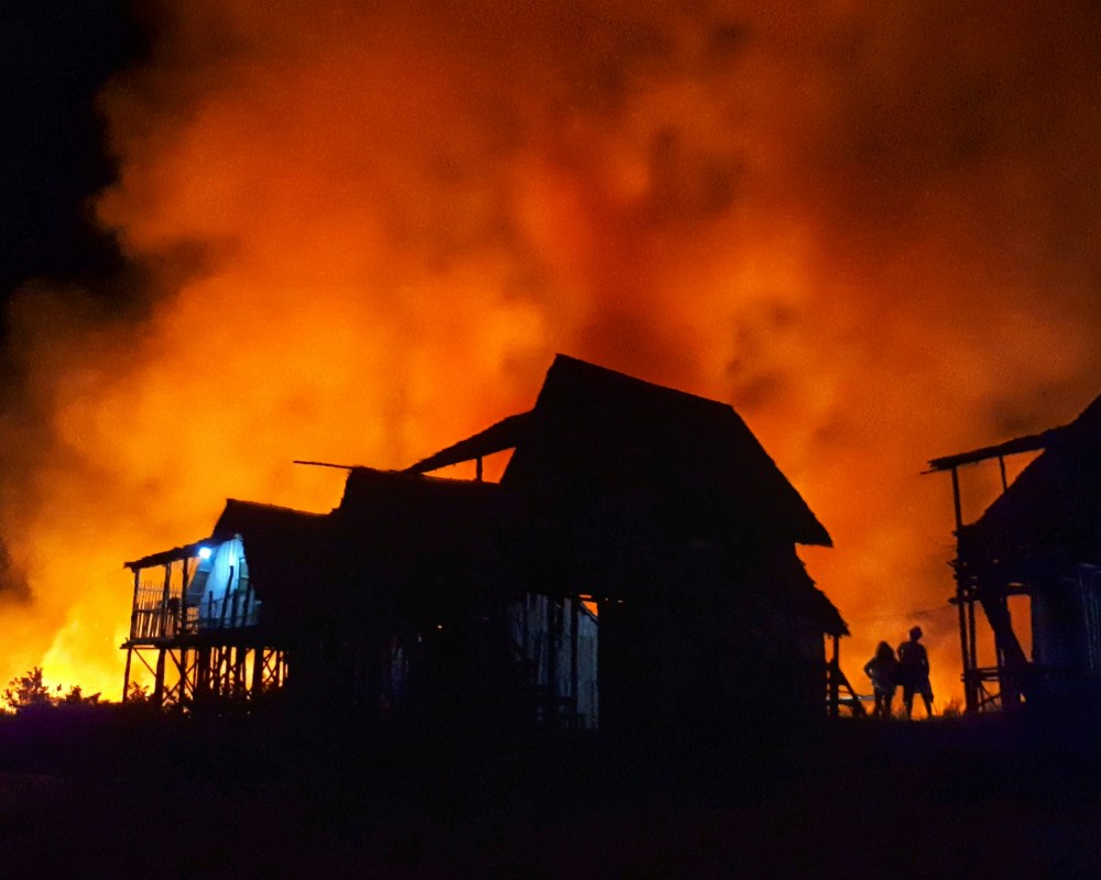 Silhouetted buildings are set against a large, intense orange fire with thick smoke filling the night sky. Two people stand near the burning structures, partially illuminated by the flames.