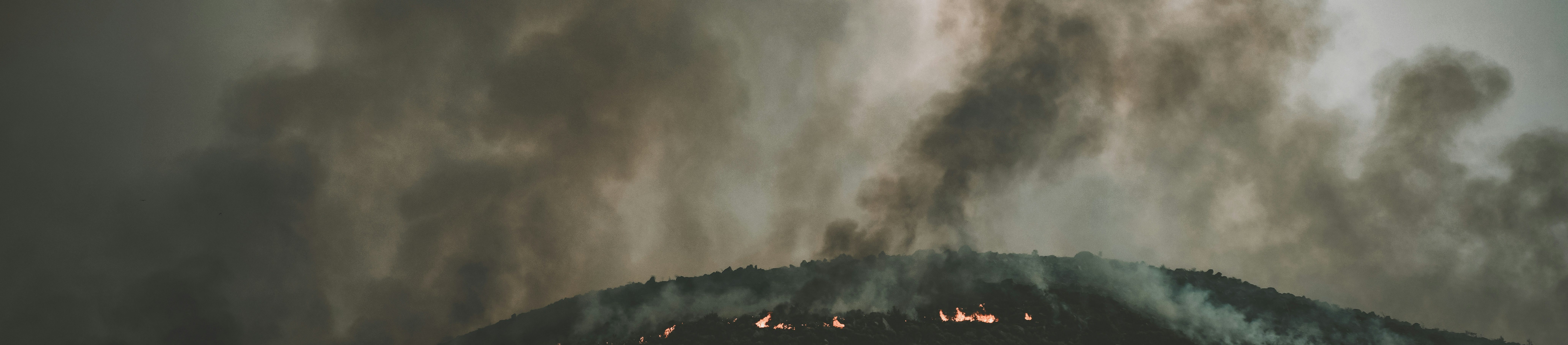 A wildfire burns along the ridge of a dark hill, with thick smoke billowing into the cloudy sky above, creating a dramatic and intense scene.